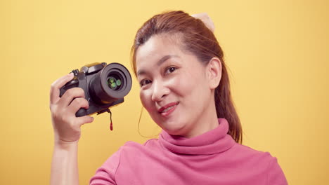 광고를 위한 카피 공간이 있는 고립된 노란색 배경에 디지털 카메라를 사용하여 분홍색 옷을 입고 노는 젊은 아시아 여성