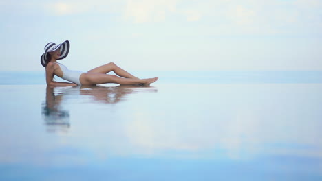 Petite-asian-female-in-a-swimsuit-and-summer-hat-lying-on-a-infinity-pool-match-up-with-tropical-sea-endless-horizon,-full-frame