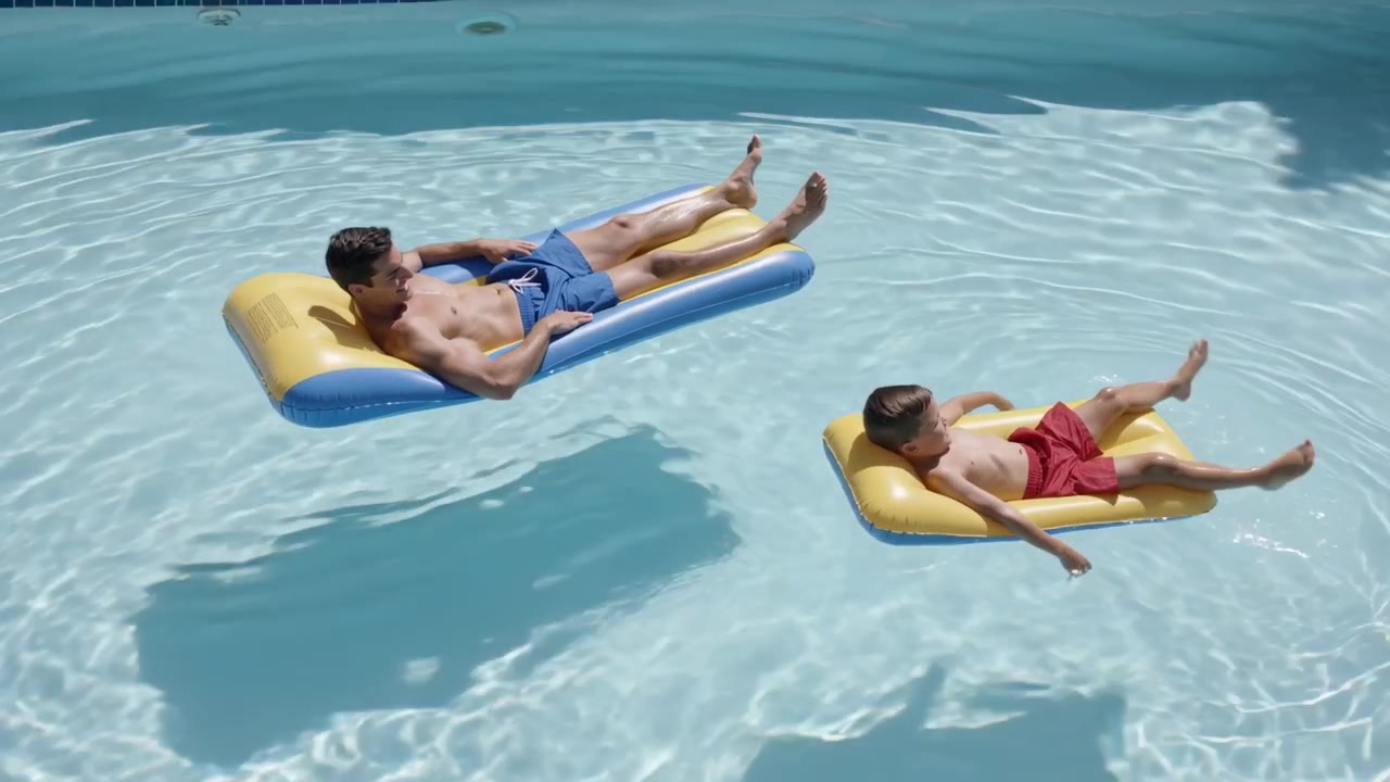 Floating man and boy relaxing with limb motion in backyard pool, wearing swim shorts, yellow floats