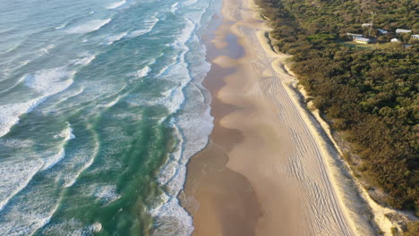 Aerial-tilt-shot-of-large-waves-hitting-the-Fraser-island-beach,-golden-hour-in-Australia
