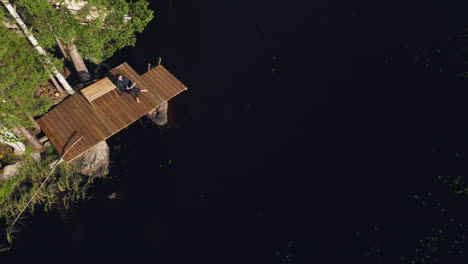 aerial descending view of a woman sitting on a deck reading a book on the water