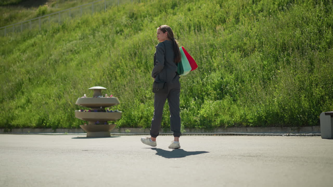 회색 옷을 입은 여자가 그녀의 뒤에 다채로운 쇼핑 가방을 가지고 고, 은 날 언덕 에서 산책하고, 배경에는 푸른색과 밝은 빛 아래의 벤치가 있습니다.