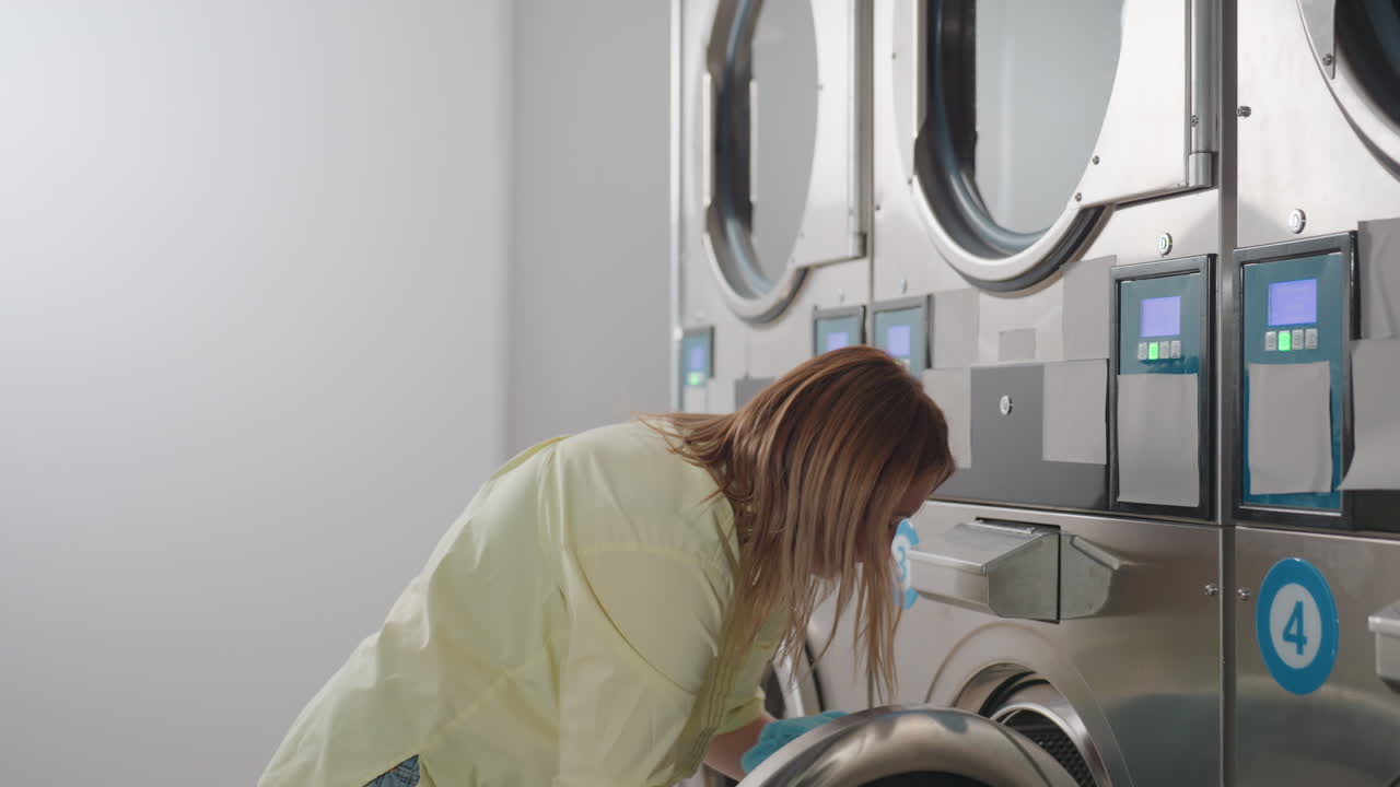 Laundry worker removes clothes from front load washer, collects textiles into basket, turns away for transport inside bright laundromat, stainless machines in row, routine hygiene service