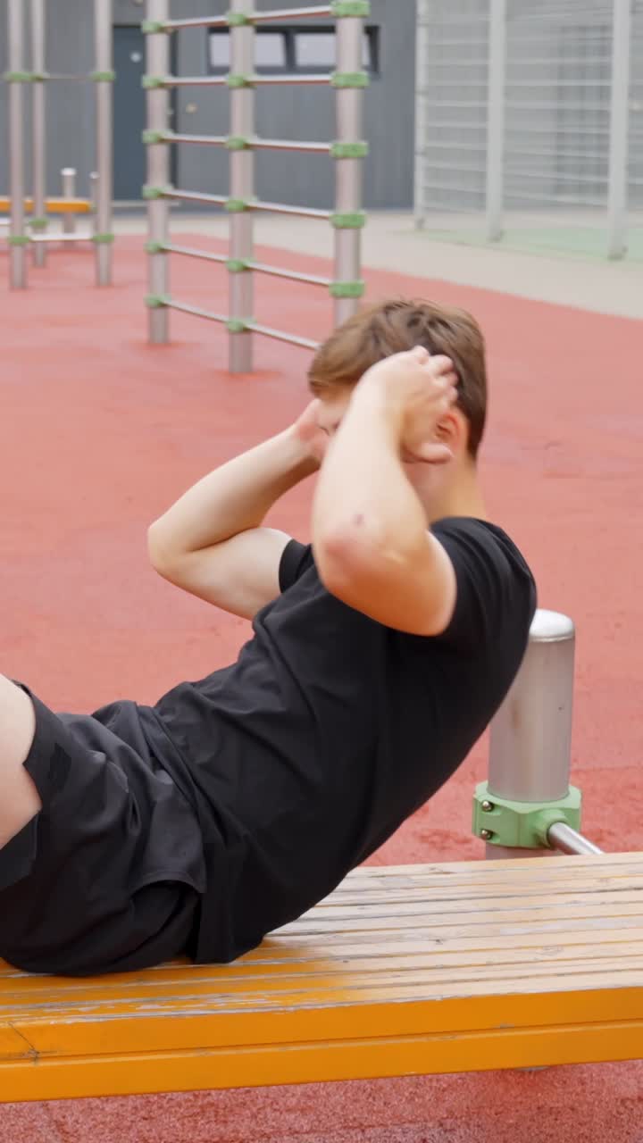 Vertical, young man in black workout clothes does crunches on exercise bench, slow motion