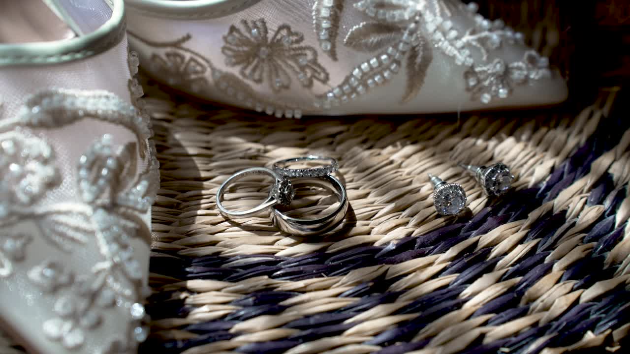 Wedding accessories are arranged on a woven mat. Shoes, rings, and earrings are clearly visible in the sunlight. The setting shows preparation for a special event