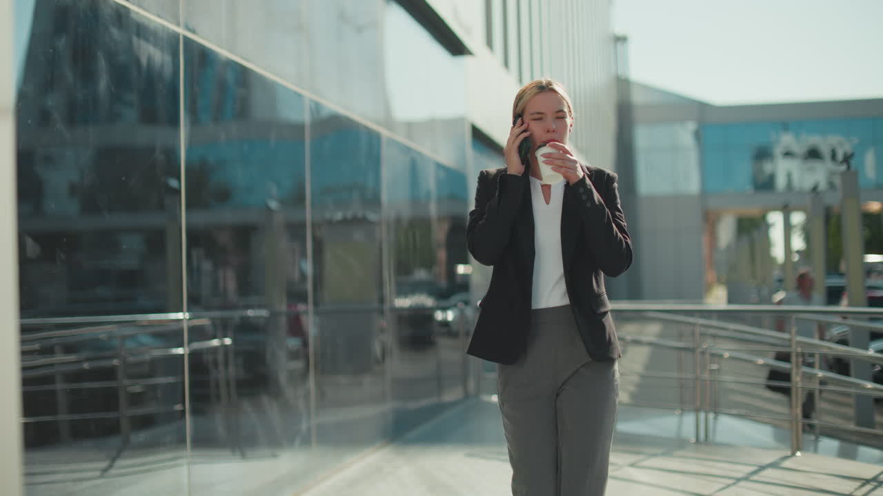 Woman walking with coffee in hand, sipping while on phone call near iron railing, modern glass building reflects parked cars, blurred figure walks behind, bright sunny day in business district