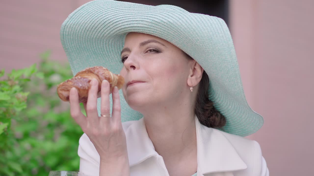 primer plano de una mujer atractiva comiendo un delicioso croissant y sonriendo a la cámara. retrato de una turista confiada descansando al aire libre. una elegante dama adulta disfrutando de un día soleado al aire abierto.