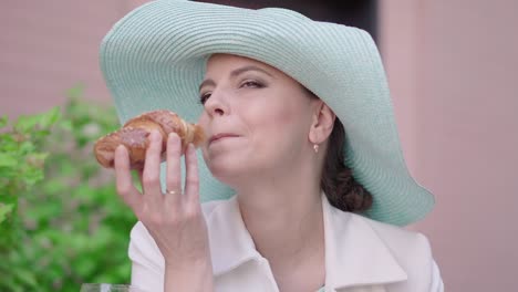 close-up of attractive woman eating delicious croissant and smiling at camera. portrait of confident female tourist resting outdoors. elegant mid-adult lady enjoying sunny day outdoors.