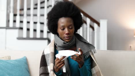 close up portrait of african american female wearing a scarf, sitting at home on a sofa and blowing hernose in a tissue