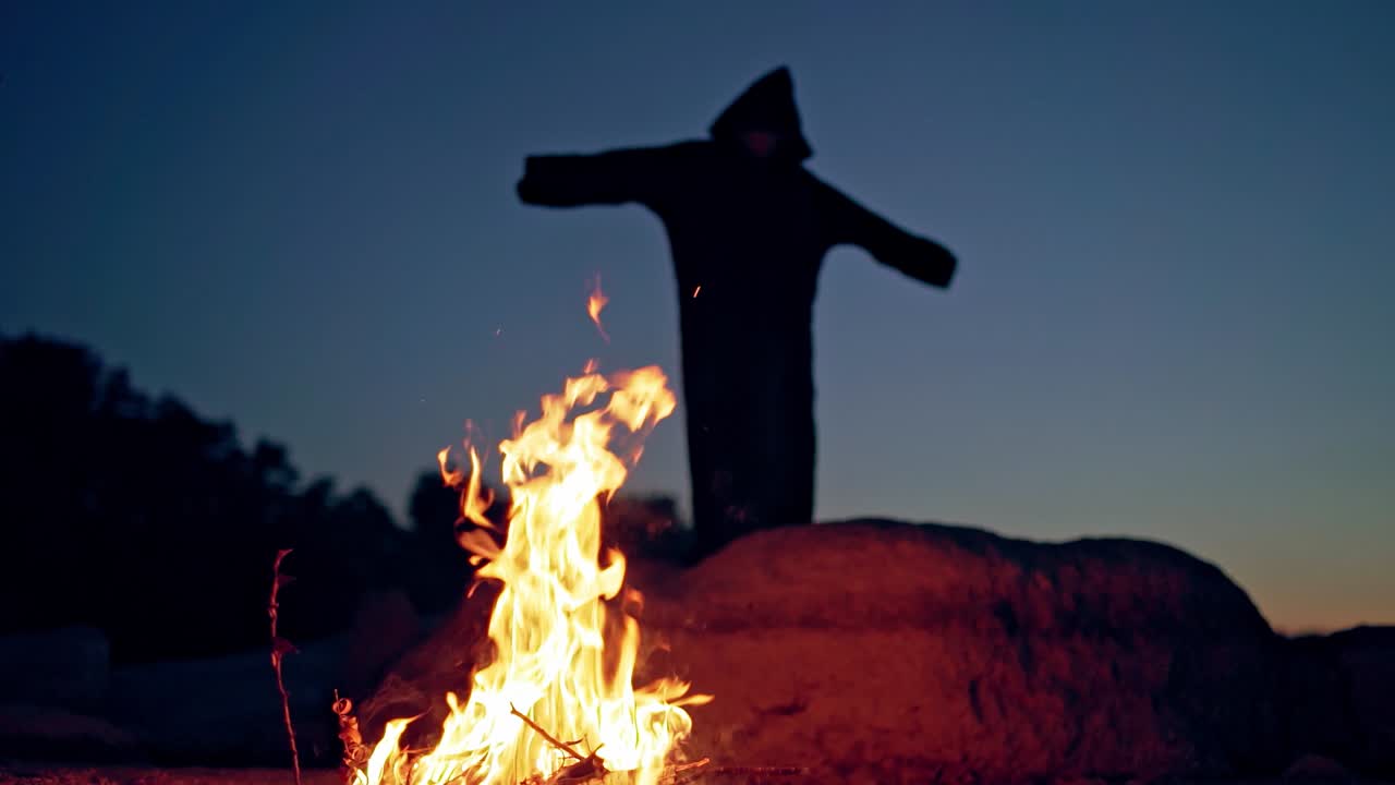 Evil witch in black cloak with hood near the fire. Halloween costume.