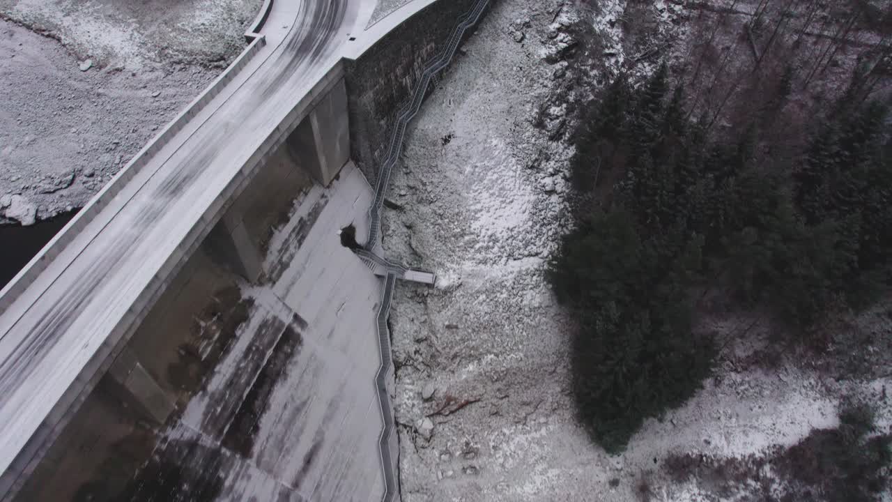 Aerial bird's eye shot of a red car passing by highway over the concrete arched dam surrounded by a mountainous terrain on a cold morning