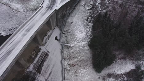 Aerial-bird's-eye-shot-of-a-red-car-passing-by-highway-over-the-concrete-arched-dam-surrounded-by-a-mountainous-terrain-on-a-cold-morning