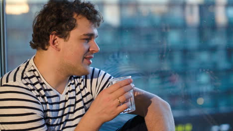 Hombre-Feliz-Sentado-Junto-A-La-Ventana-Con-Un-Vaso-De-Agua