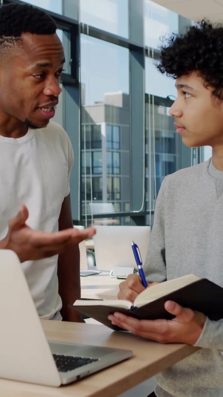 Video verticale: Un mentore che indica e guida un adolescente che scrive in uno studio con laptop, quaderno e penna blu.