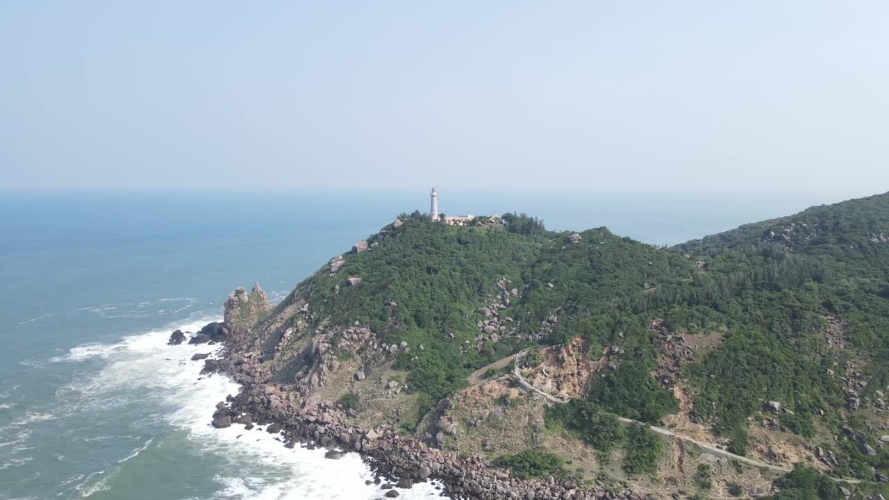 White lighthouse stands on rocky cliff above green ridge and rugged coastline, overlooking open sea and island horizon, dramatic location at Strumble Head Pembrokeshire Wales, cloudy sky adds mood