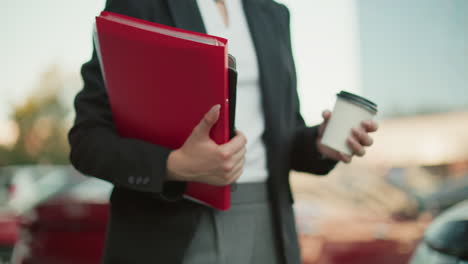 partial view of secretary in black suit holding file folder and coffee cup while walking along path with blurred background of parked cars and urban buildings