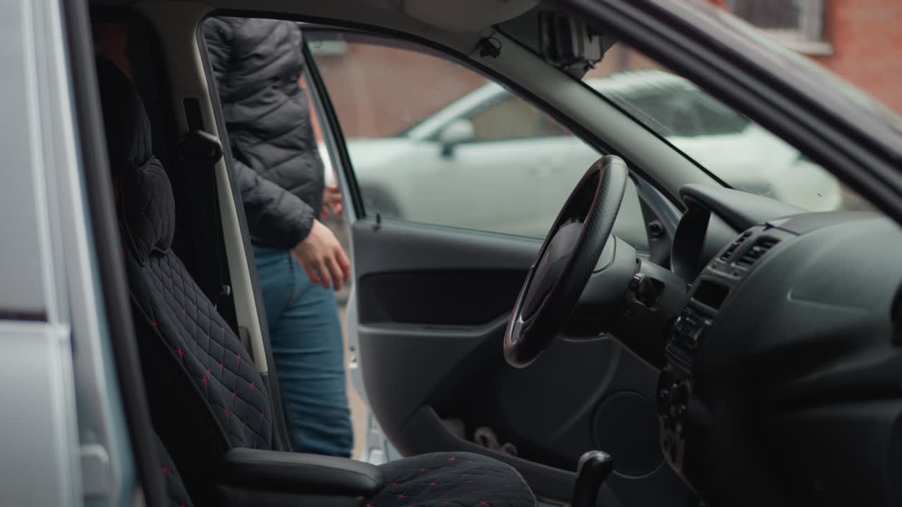 Inside view from drivers seat showing driver entering car, sitting and placing hands on steering wheel while cars parked and moving pass by window past brick building during winter commute