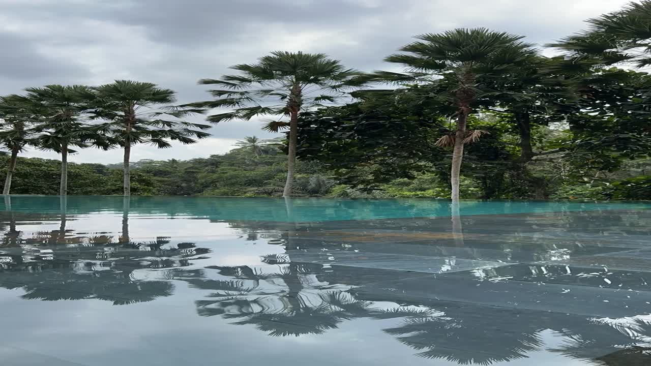 Infinity pool blends with lush tropical trees in Ubud region of Bali as calm water reflects soft sky tones creating peaceful retreat mood captured through smooth timelapse perspective