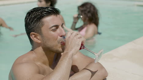 hombre guapo bebiendo un cóctel en la piscina mientras sus amigos juegan detrás