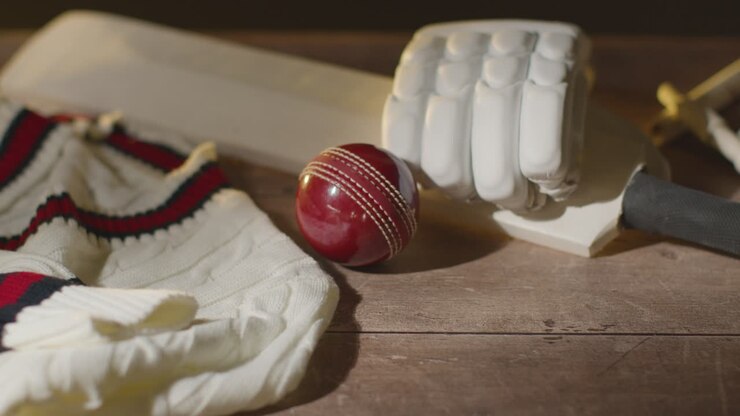Natura morta di cricket con primo piano di mazza, guanti, paletti, maglione e bails, con la palla che rotola nell'inquadratura
