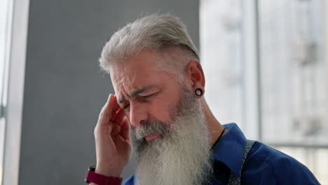 cerrar un anciano con cabello gris una barba exuberante y un anillo en la oreja en una camisa azul masajea su templo debido al dolor en la cabeza y muchos pensamientos que lo persiguen en un apartamento moderno