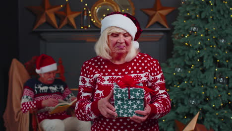 Abuela-Madura-Con-Suéter-Festivo-Presentando-Caja-De-Regalo-De-Navidad,-Sonriendo,-Mirando-A-La-Cámara