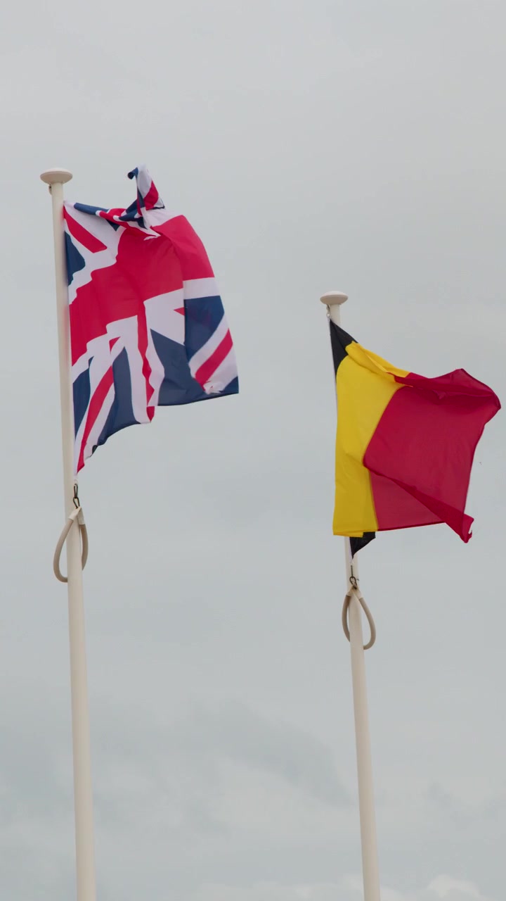 Two national flags flutter vigorously on tall poles against cloudy skies, shot with steady camera