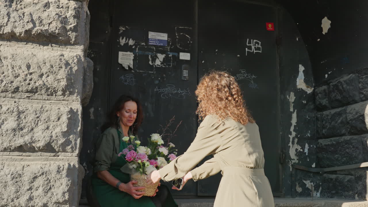 Donna che riceve un bouquet all'angolo della strada mentre una cliente consegna una composizione floreale pastello a una fiorista seduta nell'ingresso in pietra, la calda luce del sole illumina un bouquet di peonie e rose, scambio spontaneo di gentilezza, cesto.