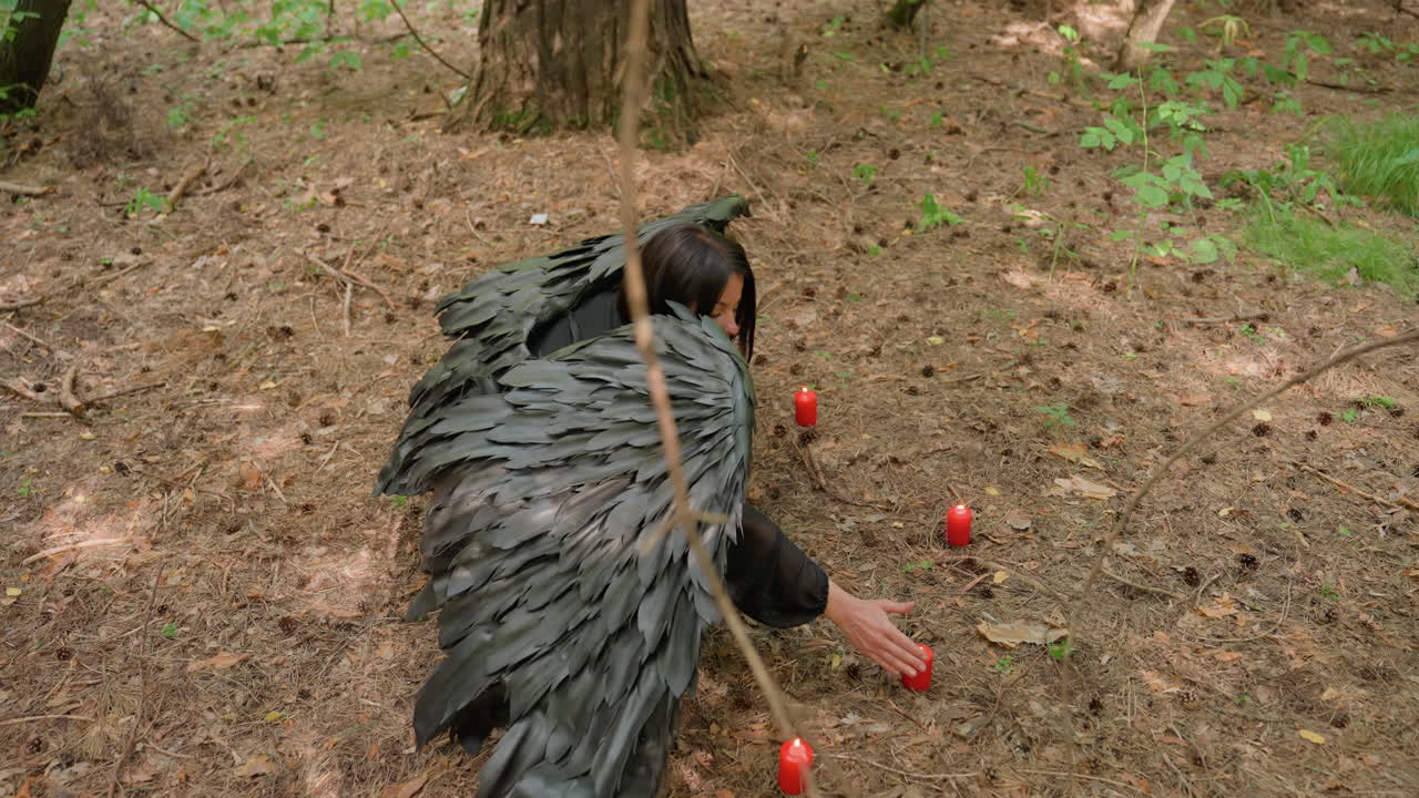Top view of black fairy with large wings kneeling in forest surrounded by glowing red candles, gently touching flame before prayer, magical and spiritual moment filled with mystery