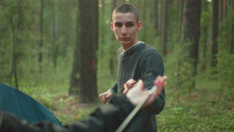 duo lovers standing in wooded forest environment fixing flexible tent pole together while preparing campsite, surrounded by green foliage and tall trees, engaging in teamwork