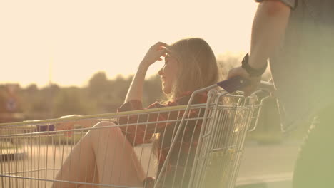 back view. cheerful young couple in love man and woman laughing and having fun while riding carts in supermarket parking in slow motion at sunset.