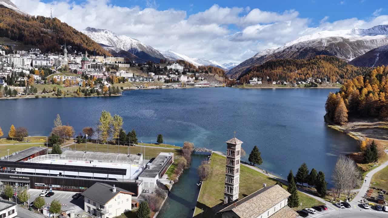 An aerial, high-angle view of the exclusive resort town Sankt Moritz Dorf in the Upper Engadin. Elegant hotels and luxury buildings are set against golden larch forests and snow-capped mountains