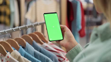 clothing store: female using smartphone with chroma key green screen display. clothes hanger with stylish branded items for retail sale in the background. close up footage of mobile device.