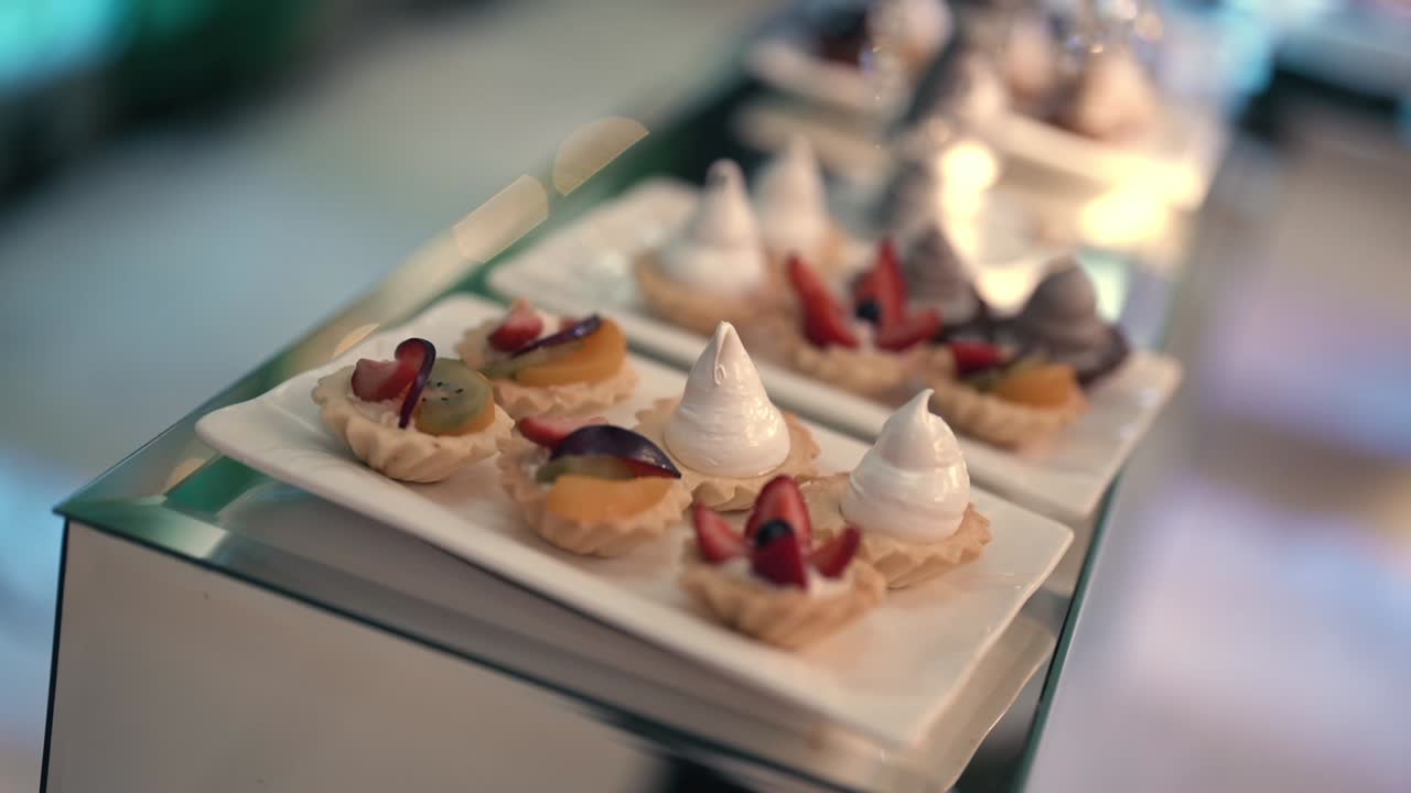 Roterende nærbillede af kamera, der fokuserer på et udvalg af gourmetdesserter ved en eksklusiv banket. Varmt omgivende lys fremhæver den sofistikerede præsentation af de søde sager.