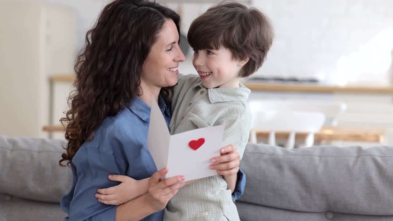 Happy mom embrace cute boy reading handmade postcard with heart for mother day or valentines holiday