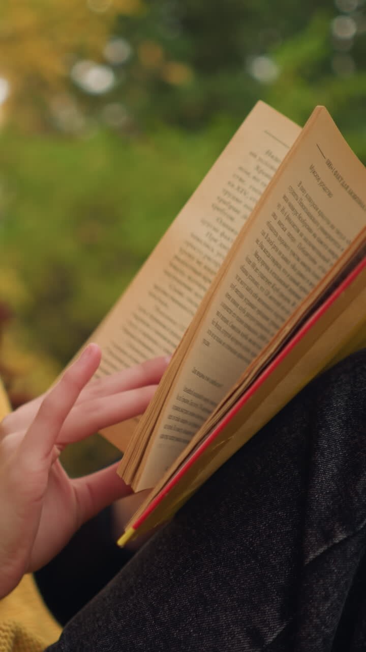 Primo piano di una donna con capelli rossi ondulati che indossa un berretto giallo e occhiali da sole, mentre sfoglia un libro nella sua mano; il viso è leggermente sfocato sullo sfondo e foglie autunnali sono sparse intorno.