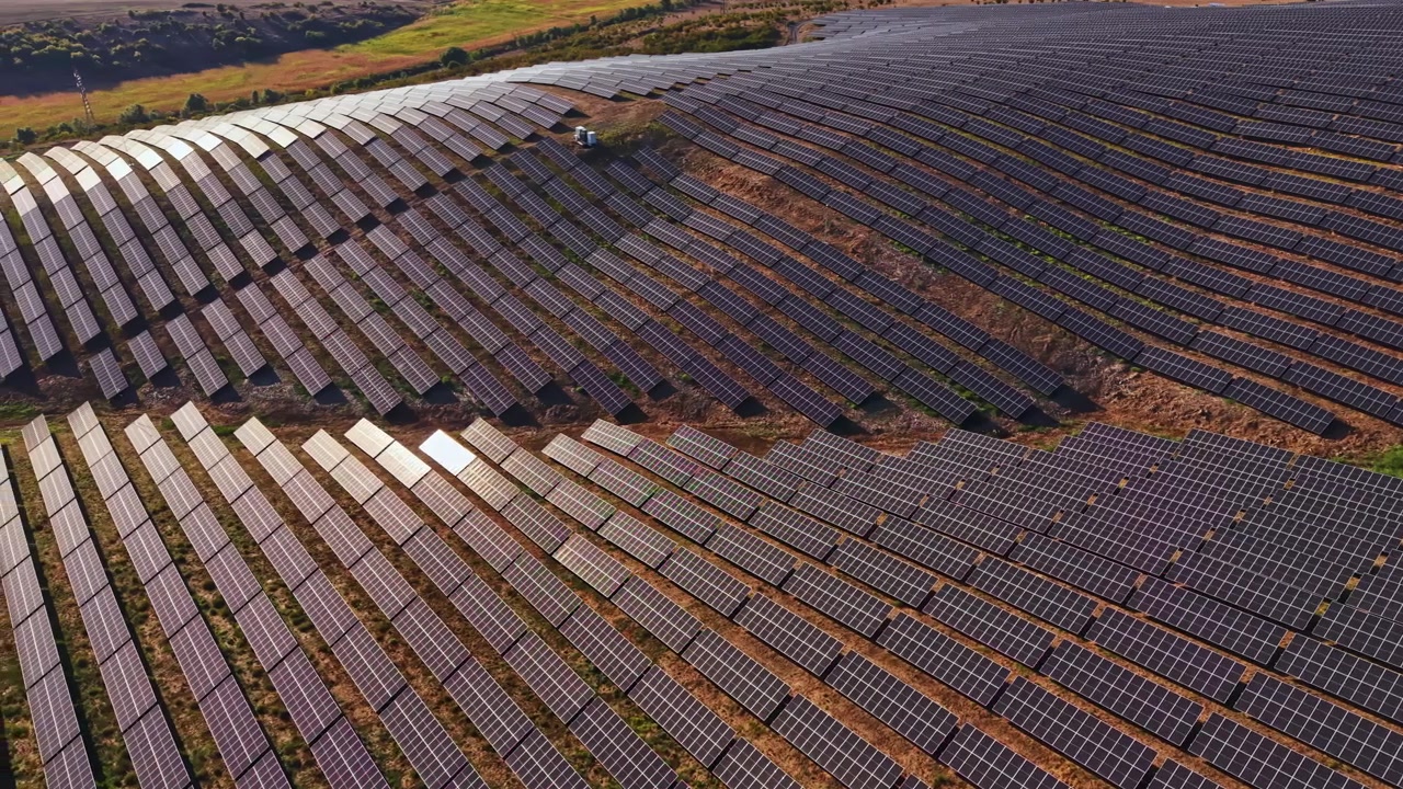Et stort solcelleanlæg dækker en bakkeskråning i et landområde i dagtimerne, med sollys der reflekteres fra panelerne og marker i baggrunden