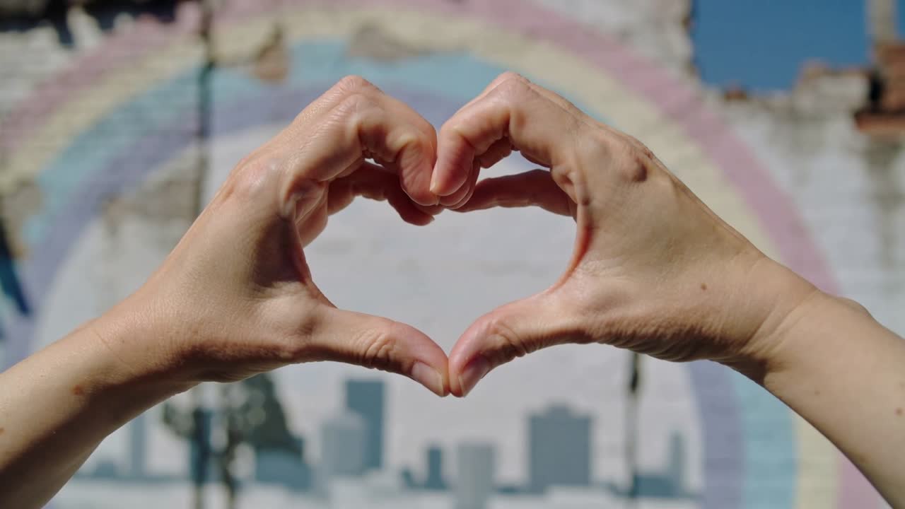 Un par de manos adultas entran en el encuadre, formando y sosteniendo un corazón frente a un mural con un arcoíris.