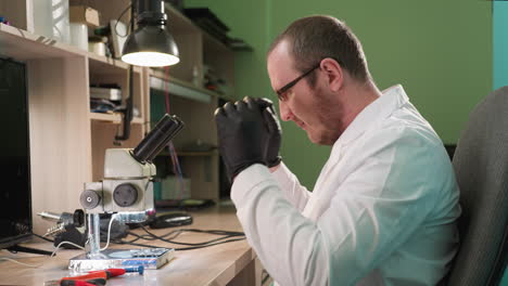 a close view of a man wearing a white lab coat inside his lab putting on his glasses while observing a circuit he is holding, the laboratory setting includes a microscope and various tools