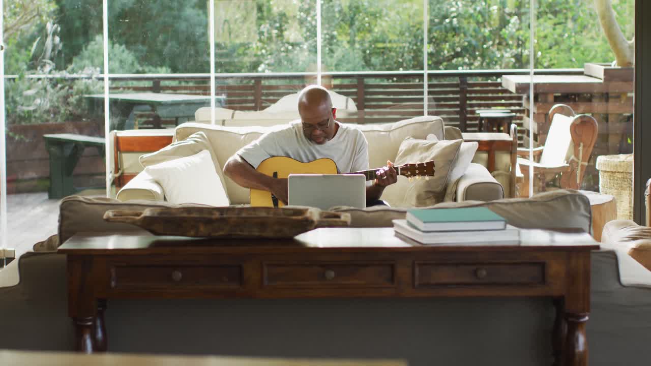 Happy african american senior man in living room playing acoustic guitar and using laptop