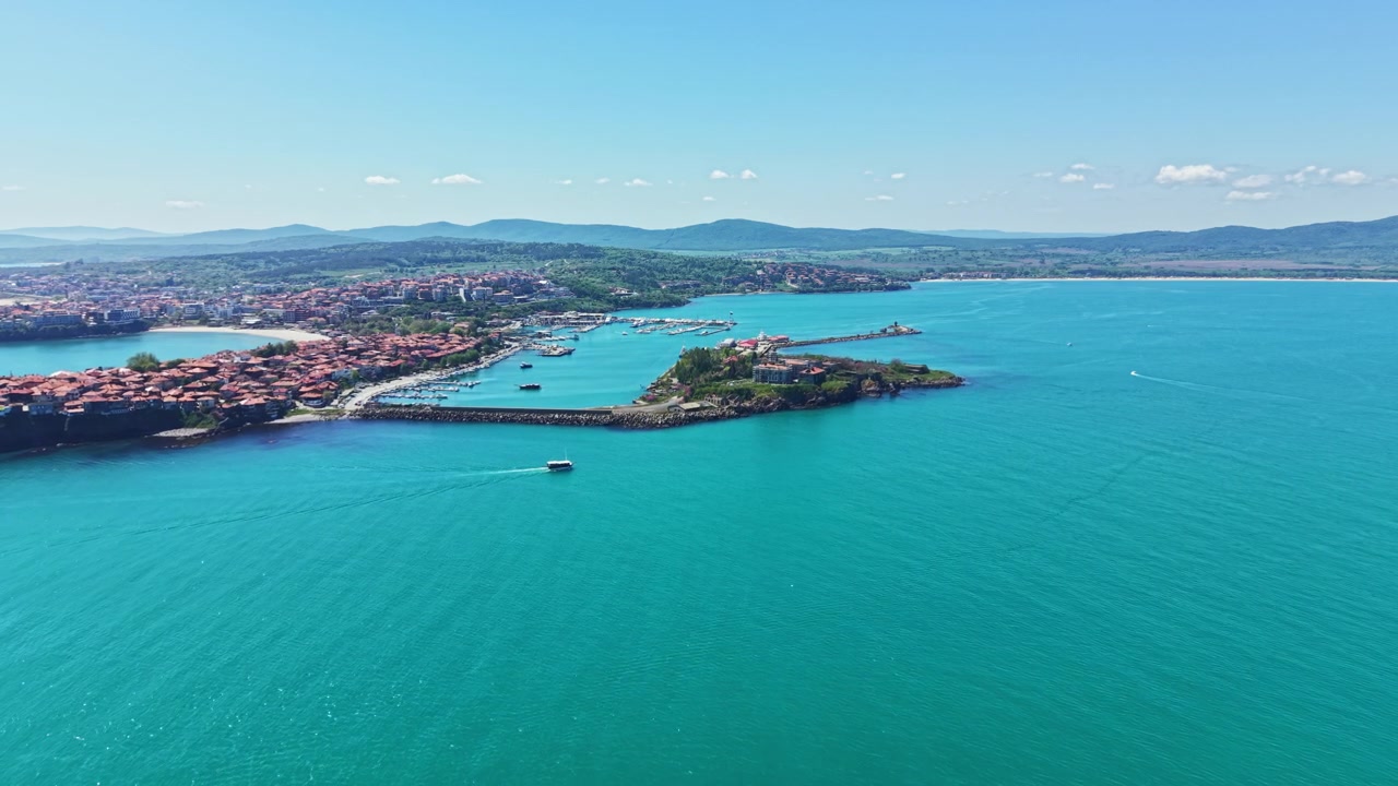 Güneşli bir günde, berrak mavi gökyüzü altında, deniz kenarında bir marina ve küçük bir kasabanın kıyı manzarası, arka planda uzaktaki tepelerle.