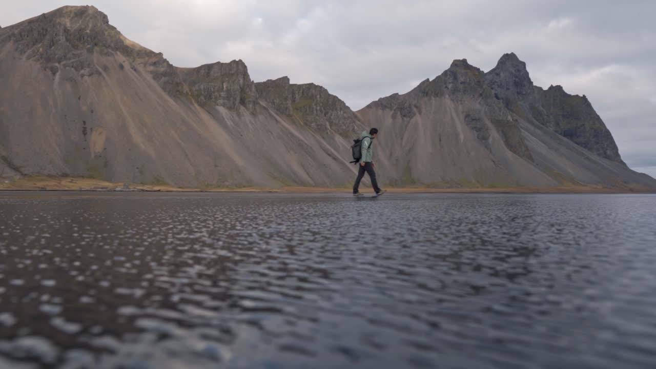 A wide shot captures a lone figure, an adventurer or photographer, walking along the dark, reflective black sand beach of Stokksnes in Iceland.