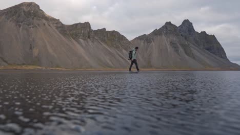a wide shot captures a lone figure, an adventurer or photographer, walking along the dark, reflective black sand beach of stokksnes in iceland.