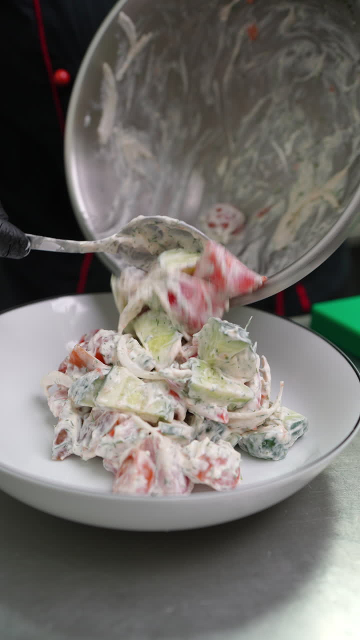 Vertical footage Professional chef wearing black uniform and gloves adding creamy white sauce to fresh vegetable salad with cucumbers, tomatoes, and onions in restaurant kitchen