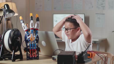 un chico asiático lindo que tiene dolor de cabeza mientras trabaja sobre una mano de cyborg en una computadora portátil en casa