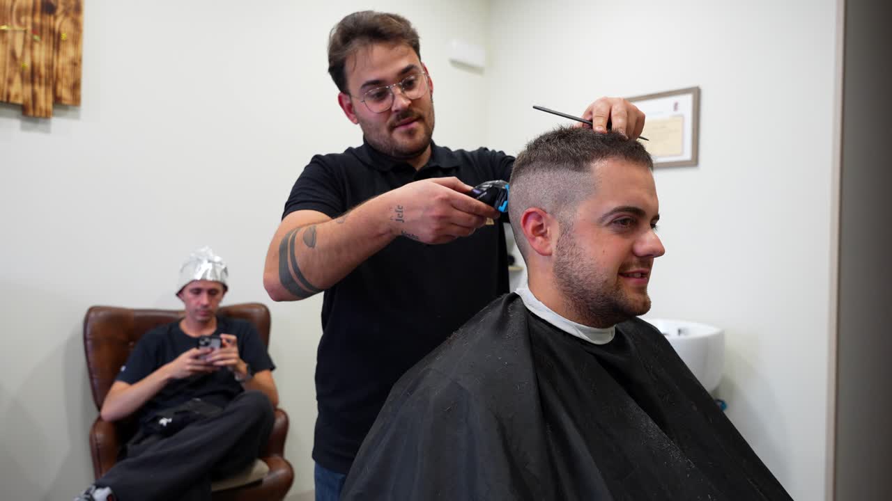 Action shot of a barber using a trimmer then scissors while a second customer waits with silver foil on his head