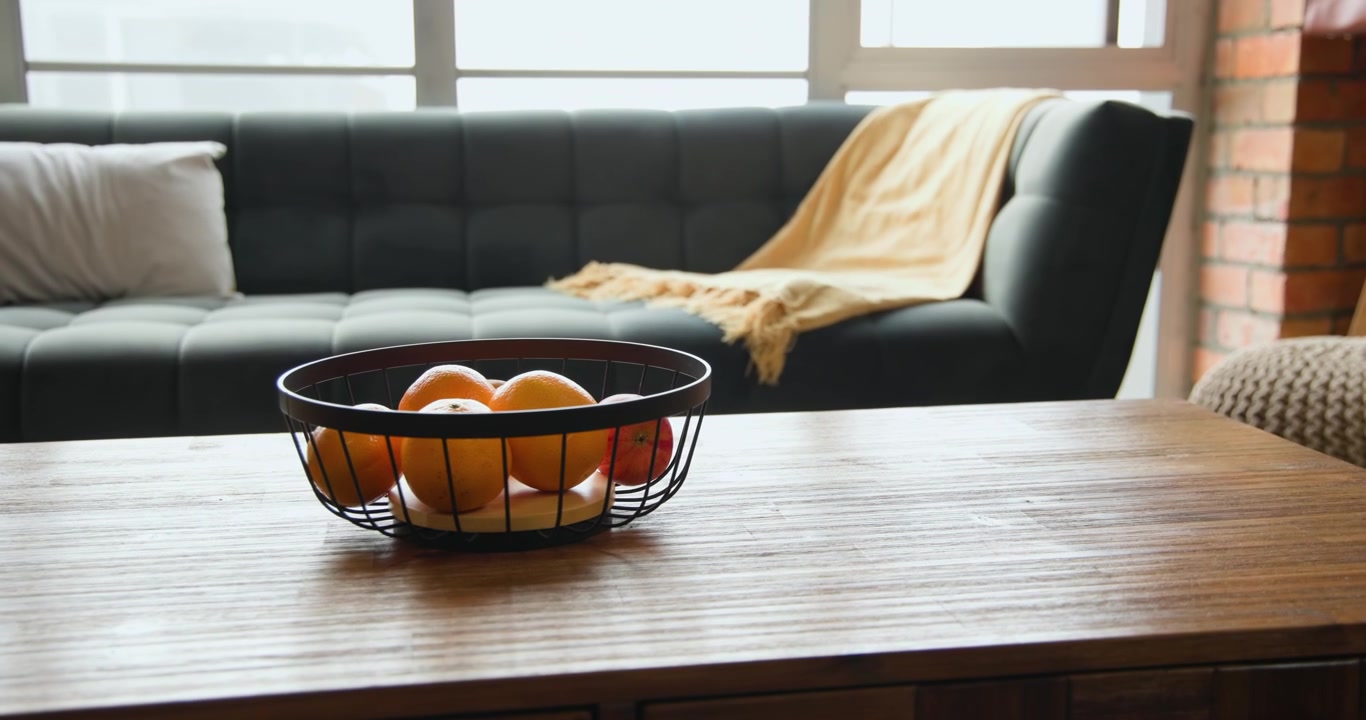 Camera is pulling back, revealing wire basket of oranges and apple on wood table, establishing calm