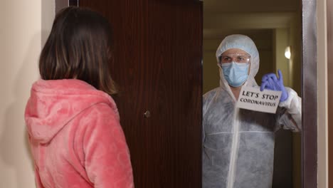 a man in a protective suit and mask holds up a sign that says 'let's stop coronavirus' to a woman wearing a face mask. they are in a hallway of a building.