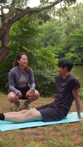 Vertical-Video-Of-Woman-Teaching-Man-Wearing-Sports-Clothing-In-Outdoor-Yoga-Class-Surrounded-By-Forest-And-Lake-Or-River-Shot-In-Real-Time