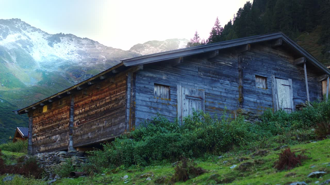 vieja cabaña alpina de madera cerca después de la puesta del sol con rayos de luz detrás y un paisaje alpino con árboles y montañas alpinas en el fondo y arbustos verdes en el frente